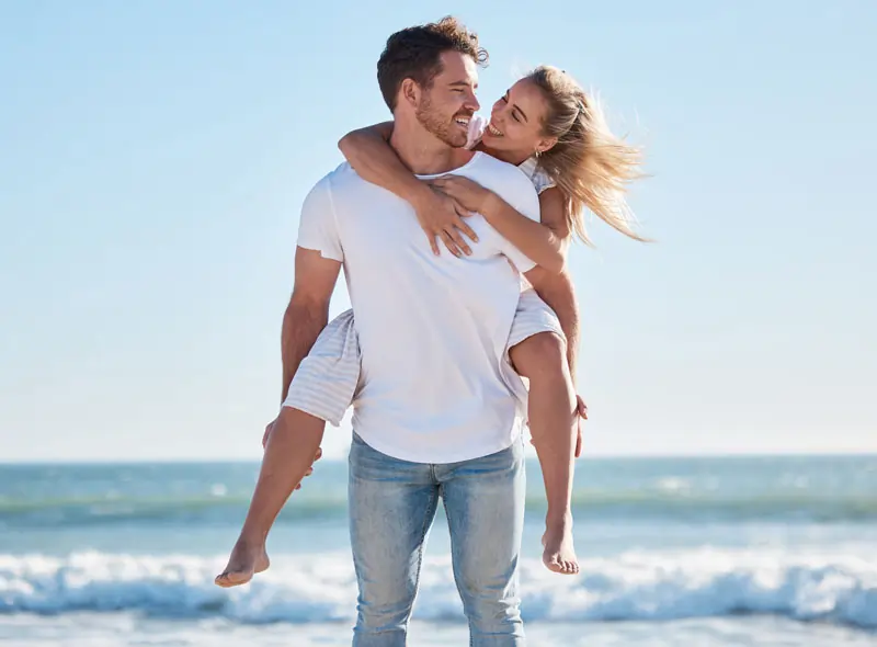 Ein glückliches Paar am Strand, der Mann trägt die Frau auf dem Rücken, symbolisiert den Wiederaufbau von Vertrauen und Selbstbewusstsein nach einer Trennung.