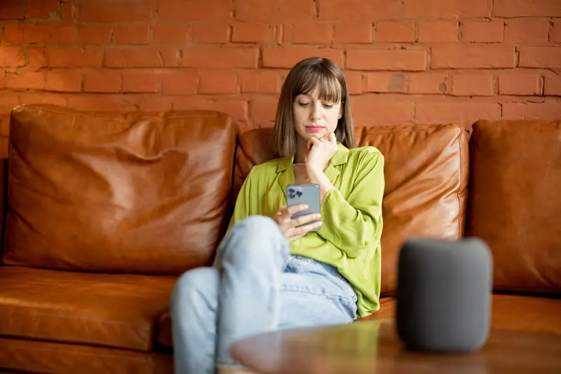 Frau sitzt nachdenklich auf dem Sofa und schaut auf ihr Handy, während sie über den Kontakt nachdenkt, obwohl er eine andere hat.