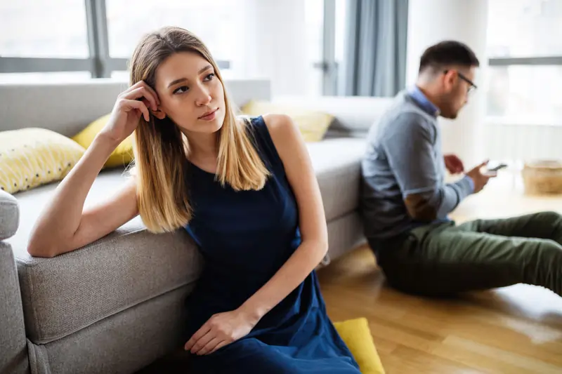 Nachdenkliche Frau sitzt auf dem Boden, während ihr Partner auf dem Handy tippt, was ein Anzeichen dafür sein könnte, dass sie das Gefühl hat, er hat eine andere.