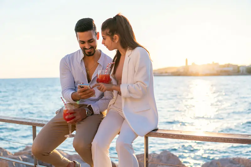 Lachendes Paar sitzt mit Cocktails am Meer und schaut gemeinsam auf ein Smartphone – zeigt die unbeschwerte Zeit in der Verliebtheitsphase einer neuen Beziehung.